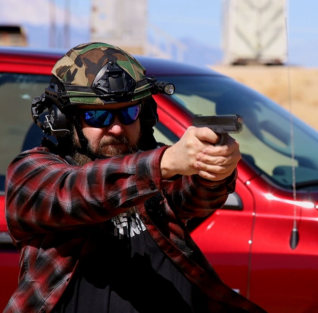 Shawn Herrin firing a handgun beside a red vehicle during an outdoor range stage